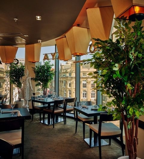 Chaophraya_Elegant_Dining_Indoor_View Interior of Chaophraya restaurant with elegant dining setup, modern wooden tables and chairs, hanging geometric lamps, and large windows overlooking city buildings.