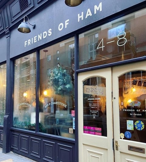 Friends of Ham Leeds City Bar Exterior Charcuterie Exterior view of Friends of Ham in Leeds City, showcasing the entrance with a beige door and large windows, the name of the establishment in large white letters, and a sign indicating it is a bar and charcuterie.