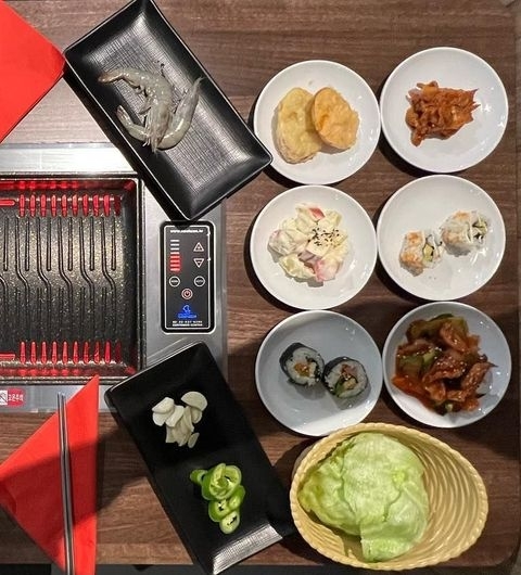 Hama Korean BBQ Buffet Assorted Cuisine Spread Assorted Korean dishes on a table at Hama Korean BBQ Buffet, including a grill with raw shrimp, small plates with dumplings, kimchi, salad, sushi rolls, spicy chicken, and a basket of lettuce leaves.