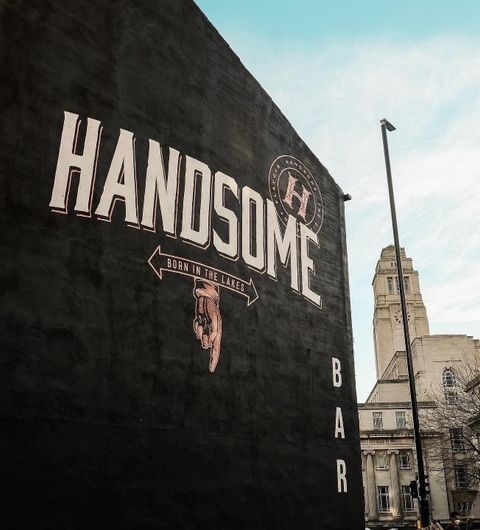 Handsome Bar Urban Wall Signage Exterior Exterior wall of Handsome Bar with bold white lettering on a dark background, featuring a logo with the text "Born in the Lakes" and a clear sky above.