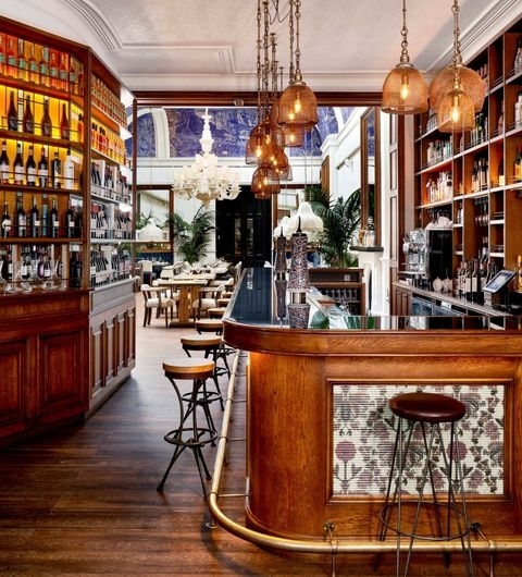 Ibérica Elegant Bar Interior Vintage Decor Elegant interior of Ibérica featuring a curved wooden bar with stools, pendant lights, and shelves lined with bottles, leading to a dining area with tables and chairs.