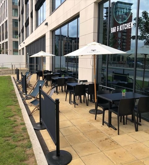 MadFransBar_OutdoorPatio_Umbrellas_DiningArea Outdoor seating area of Mad Frans Bar with black chairs and tables under white umbrellas on a paved patio, adjacent to a grassy area, with the bar's sign visible on the building's window.
