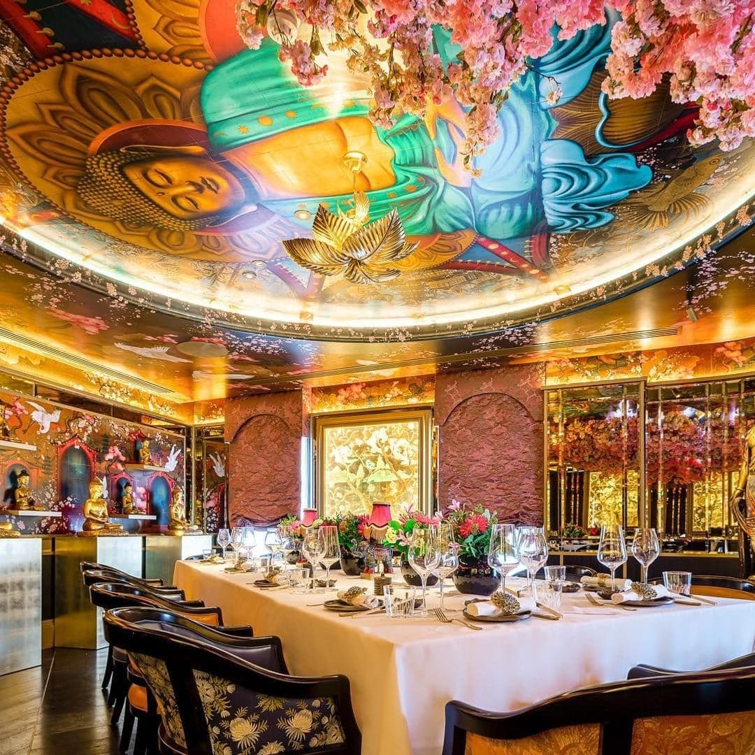 Elegant dining setup at The Ivy Asia with a vibrant Buddha ceiling mural, floral arrangements, and ornate decor.
