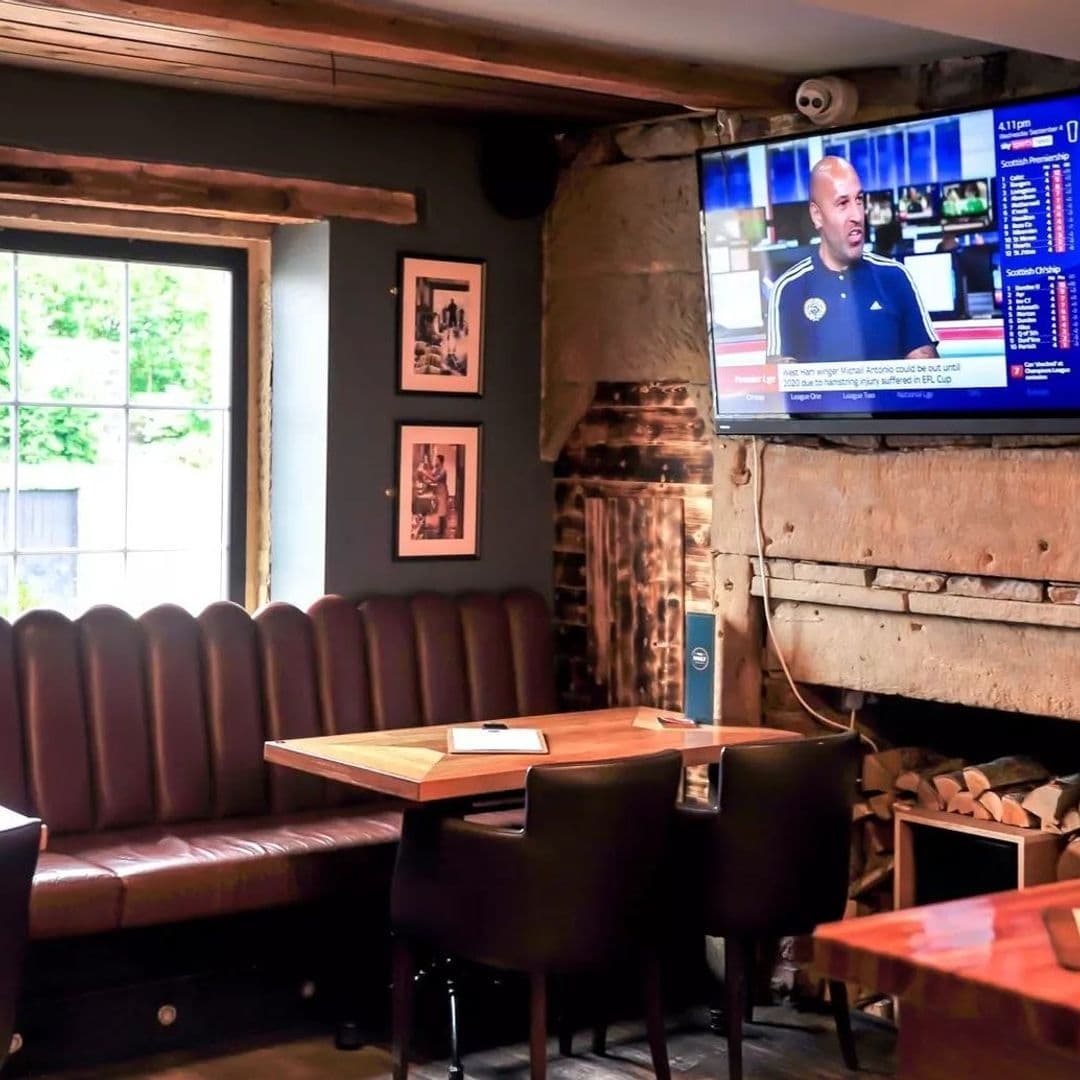 Interior of The Malt Brewhouse showing a cozy seating area with a brown leather sofa and wooden tables, framed pictures on a dark wall, and a television displaying sports. A window provides natural light, and a stack of firewood adds to the rustic ambiance.