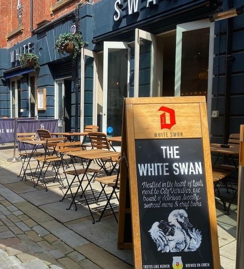The White Swan   Outdoor Seating Pub Entrance Exterior view of The White Swan pub with outdoor seating and a promotional signboard featuring the pub's name and offerings.