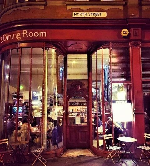 Threes A Crowd Corner Bistro Evening Dining Street View Corner view of Three’s A Crowd restaurant on North Street with illuminated interior, patrons dining inside, and outdoor seating available.