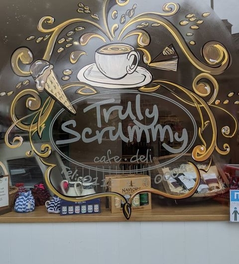 Truly Scrummy Cafe Deli Artistic Window Design Window display of Truly Scrummy cafe-deli featuring artistic gold and brown drawings of a coffee cup, an ice cream cone, and a chocolate bar, with the name 'Truly Scrummy' prominently displayed in the center.