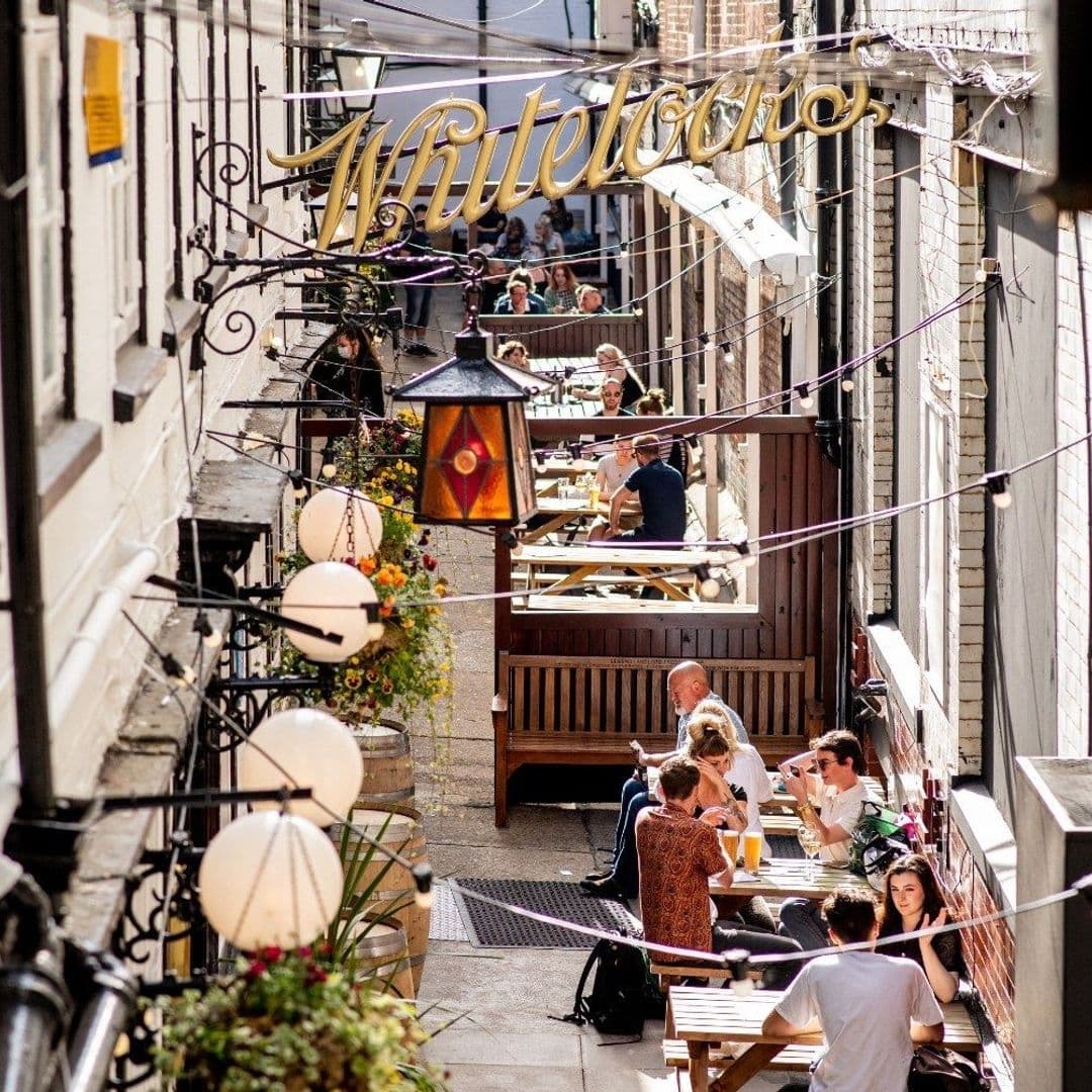 Outdoor seating at Whitelock's Ale House with patrons enjoying drinks and conversation in a narrow, sunlit alleyway adorned with hanging lanterns and floral decorations.