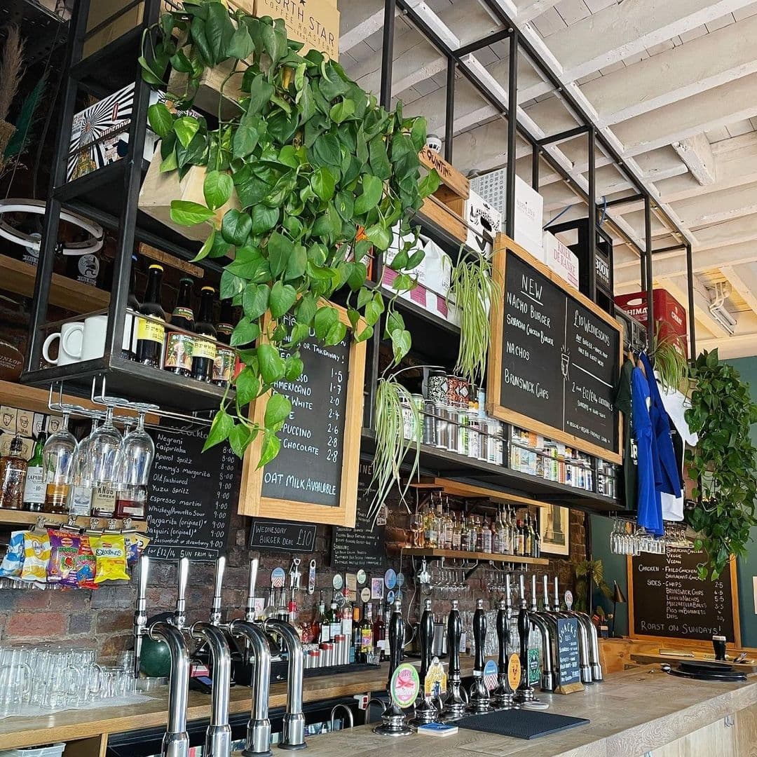 Interior view of The Brunswick featuring a bar with beer taps, a variety of spirits on shelves, and a chalkboard menu. Green plants add a fresh touch to the cozy atmosphere.