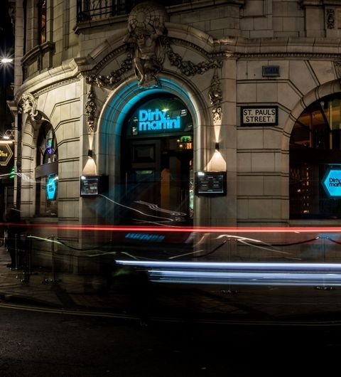 Dirty_Martini_Nighttime_Entry_Light_Trails Entrance of Dirty Martini bar at night with illuminated blue signage above the door, flanked by hanging lanterns, and a light trail from a passing vehicle in the foreground.