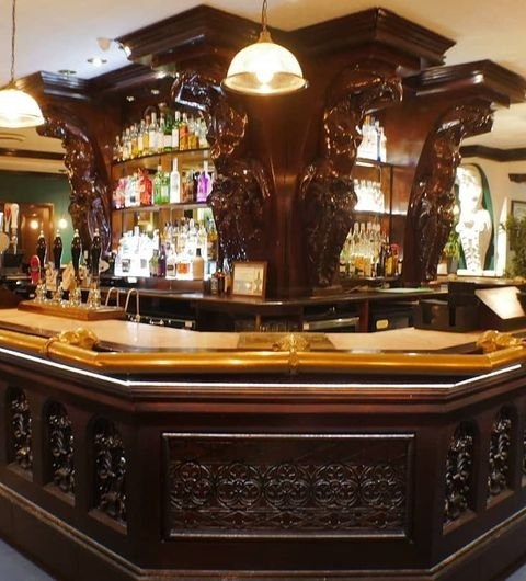 The New Conservatory Elegant Bar Interior Wood Design Elegant wooden bar at The New Conservatory with ornate carvings and a selection of beverages on shelves, illuminated by pendant lights.