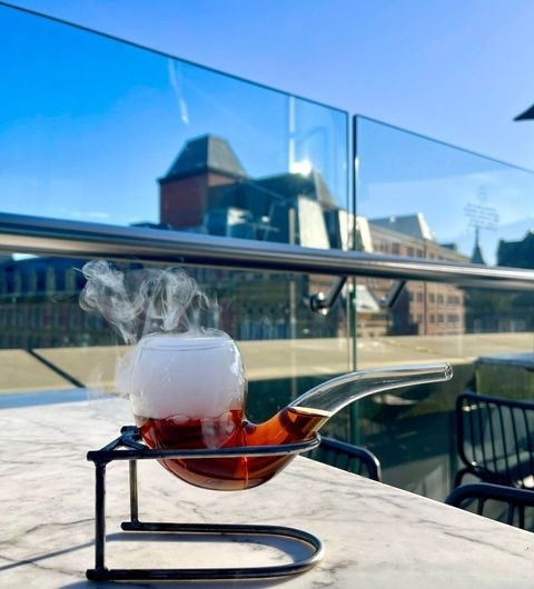 The Alchemist Trinity_Smoking Cocktail Outdoor Terrace "Unique cocktail served in a pipe-shaped glass with smoke rising from it, on an outdoor table at The Alchemist Trinity, with a clear blue sky and modern architecture in the background."