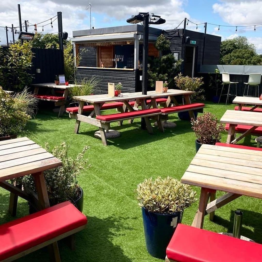 Outdoor seating area at The Woods with red cushioned picnic benches on artificial grass, surrounded by potted plants, under a string of lights with a bar cabin in the background.
