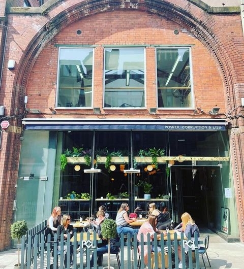 Power_Corruption_Lies_Outdoor_Dining_Brick_Architecture Exterior view of Power, Corruption & Lies restaurant with patrons dining at outdoor tables under a sunny sky, surrounded by green potted plants and the establishment's name displayed above the entrance.