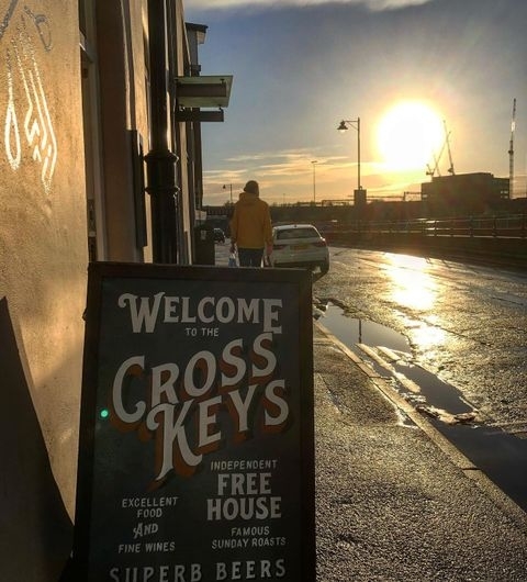 The Cross Keys Sunset Signage Streetview Sunset view outside The Cross Keys with a prominent signboard featuring the pub's name, advertising excellent food, fine wines, and superb beers, with a person walking away in the background.