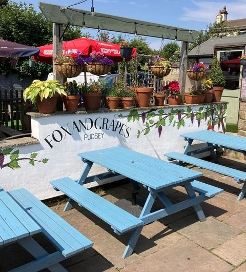 Fox and Grapes Pudsey Sunny Patio Benches Floral Decor Sunny outdoor seating area at Fox & Grapes – Pudsey with blue picnic benches, colorful hanging flower baskets, and branded umbrellas.