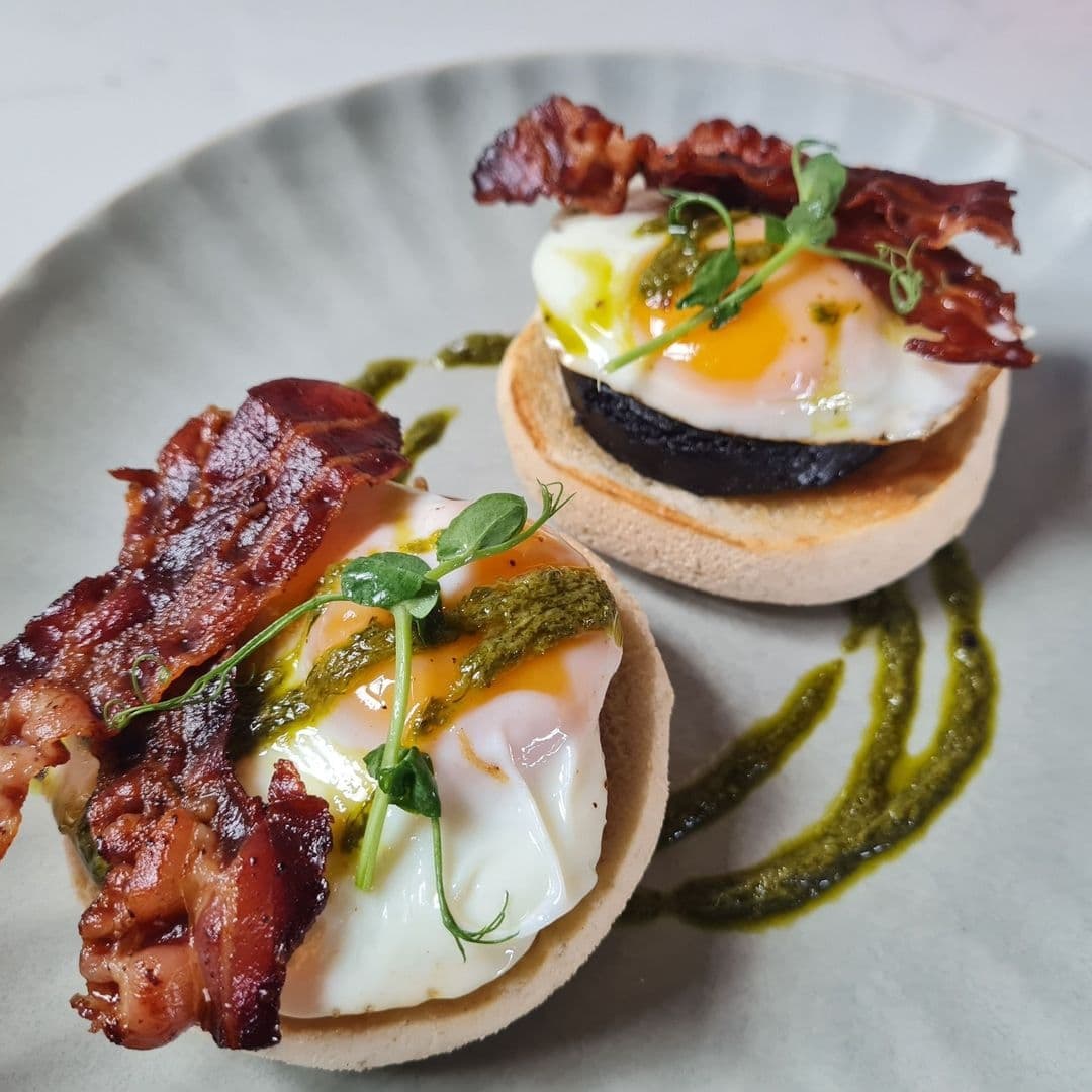 Alt text: "Delicious breakfast at Pooky’s Deli & Cafe featuring two English muffins topped with crispy bacon, sunny-side-up eggs, and a drizzle of vibrant green pesto, served on a modern grey plate."