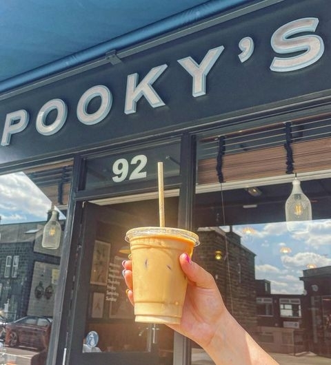 Pookys Deli Cafe Iced Coffee Exterior Sunny Day Hand holding an iced coffee in front of Pooky’s Deli & Cafe with clear blue skies in the background.