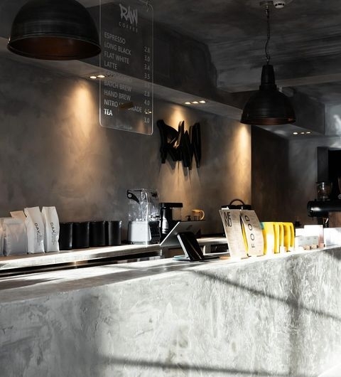 Raw_Coffee_Cafe_Interior_Modern_Industrial_Style Interior of Raw Coffee shop with a modern, industrial design featuring a concrete counter, black pendant lights, and a menu board displaying coffee options. Bags of coffee and a coffee machine are visible on the counter.