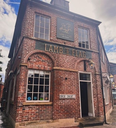 Lamb_And_Flag_Historic_Pub_Brick_Building Exterior view of the Lamb and Flag, a traditional brick building with a signboard, arched windows, and a white door, located on a cobbled street.