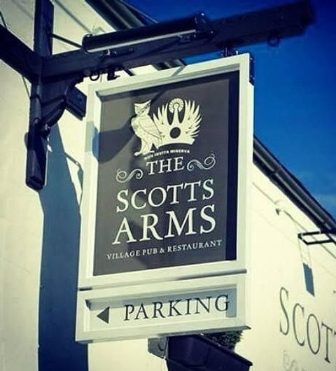 ScottsArms PubSign VillageRestaurant Parking Hanging sign for The Scotts Arms, a village pub and restaurant, with a parking sign below it, against a white building facade under a clear sky.