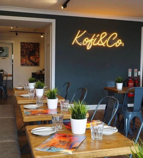Kofi Co Wetherby cozy restaurant interior modern dining ambiance Alt text: Interior of Kofi&Co – Wetherby, featuring wooden tables set with plates and glasses, small potted plants, and a neon sign on the wall. The space is inviting and modern, with artwork visible in the background.