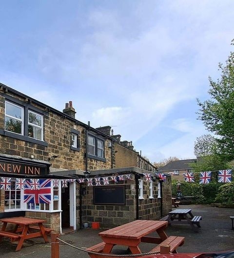 New Inn Yeadon pub exterior UK flags outdoor seating Alt text: Exterior view of the New Inn – Yeadon, featuring a stone building with British flags hanging across the outdoor seating area. The scene includes wooden picnic tables and greenery in the background under a partly cloudy sky.