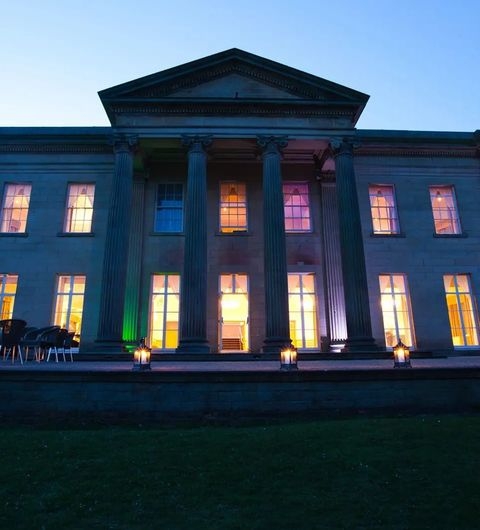 The Mansion elegant architecture evening lights historic building The Mansion, a grand building featuring tall columns and large windows, illuminated at dusk. The facade showcases a classic architectural style, with colorful lights enhancing its beauty. A well-maintained lawn and outdoor seating are visible in front of the structure.