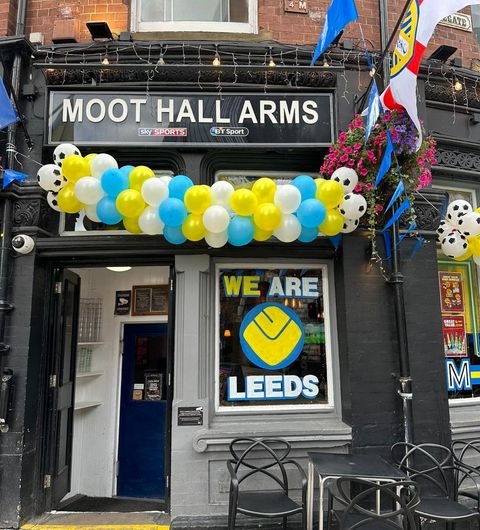 The Moot Hall Arms leeds pub decorations balloons sports fans Alt text: The Moot Hall Arms pub featuring a colorful balloon arch in blue, yellow, and white. A sign in the window reads "WE ARE LEEDS," and there are outdoor seating chairs in front of the entrance.