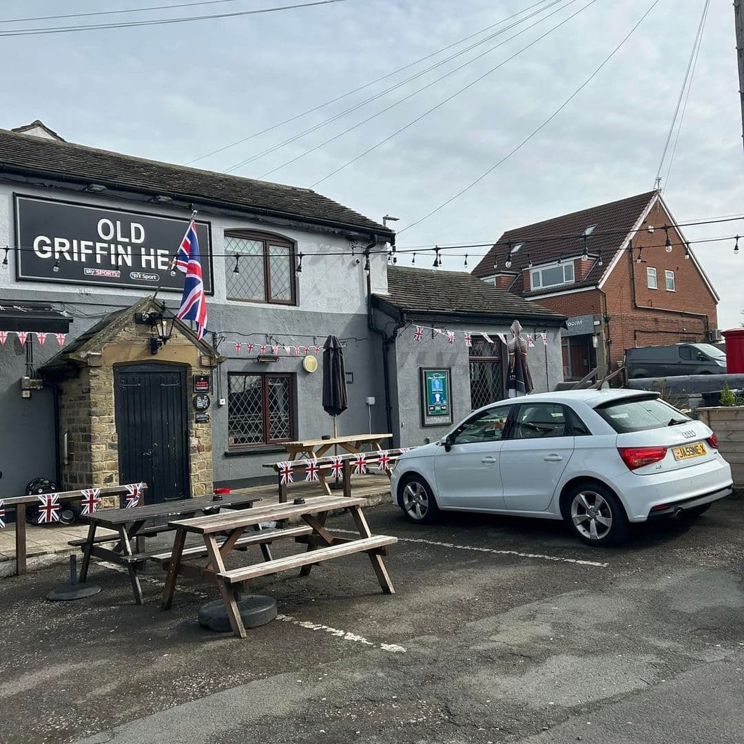 Alt text: Exterior view of The Old Griffin Head pub, featuring a British flag, outdoor seating, and a parked white car. The building has a stone entrance and decorative bunting, set against a cloudy sky.