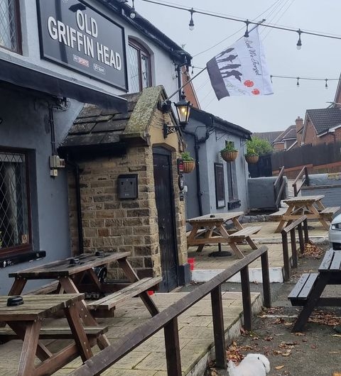 The Old Griffin Head pub exterior outdoor seating tourism Exterior view of The Old Griffin Head pub, featuring a stone entrance, wooden picnic tables, and hanging flower pots. A flag is visible above the entrance, and the surrounding area is quiet with a few houses in the background.