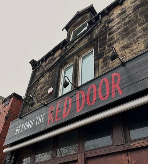 Beyond The Red Door urban architecture sign historical charm Sign for "Beyond The Red Door" featuring bold red lettering against a dark wooden background, located above a storefront. The building has large windows and a traditional architectural style, with a cloudy sky in the background.