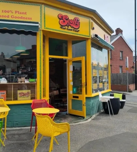 Scoffs Feel Good Food plant based restaurant colorful exterior outdoor seating Brightly colored exterior of Scoffs Feel Good Food, featuring a yellow facade and large windows. Outdoor seating includes red and yellow chairs at small tables. The entrance is welcoming, showcasing the plant-based menu.