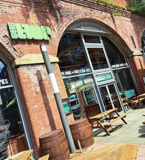 The Hop restaurant outdoor seating brick architecture urban Exterior view of The Hop, a vibrant venue with a brick facade. The entrance features large windows and a green sign. Wooden benches and barrels are placed outside, inviting guests to enjoy the outdoor seating area.
