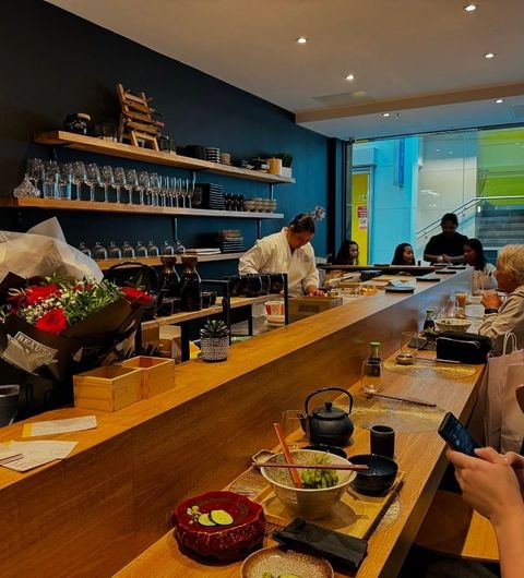 Fern Modern Sushi dining experience sushi chef ambiance restaurant interior Alt text: Interior view of Fern Modern Sushi, featuring a wooden counter with customers seated. A chef prepares sushi in the background, while shelves display glassware and bottles. Fresh flowers and dishes are on the counter, creating a vibrant dining atmosphere.