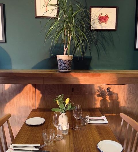 BavetteBistro cozy dining table decor plant interior A cozy dining table set for two at Bavette Bistro, featuring a wooden table with plates, glasses, and a small vase of flowers. A potted plant sits on a shelf above the table, with artwork on the wall in the background. The warm lighting creates an inviting atmosphere.