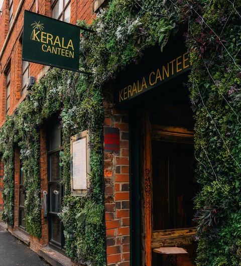 Kerala canteen greenery exterior restaurant urban Alt text: The entrance of Kerala Canteen, featuring a brick wall adorned with lush greenery and plants. A sign above the door displays the name "Kerala Canteen" in bold letters.