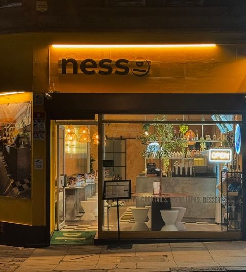 Nesso Coffee vibrant cafe exterior open sign nighttime Alt text: Exterior view of Nesso Coffee at night, featuring a bright yellow facade with the logo "ness" illuminated. The window displays a cozy interior with seating and decorative plants, and an "OPEN" sign is visible.