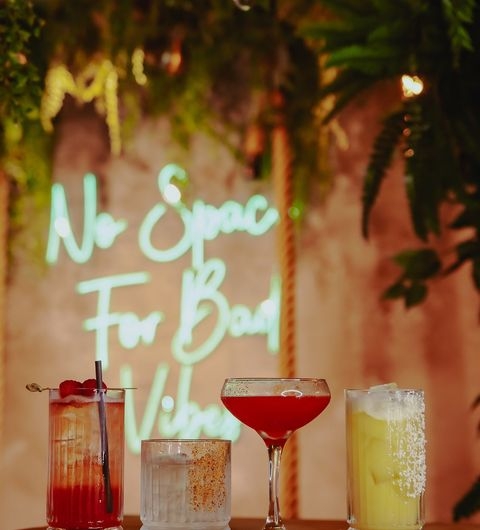 Nyla s cocktails vibrant drinks nightlife relaxation Four colorful cocktails are displayed in front of a neon sign that reads "No Space For Bad Vibes." The drinks include a red cocktail, a clear drink with a garnish, a pink cocktail in a coupe glass, and a yellow drink with bubbles. Lush greenery is visible in the background. This image captures the vibrant atmosphere at Nyla’s.