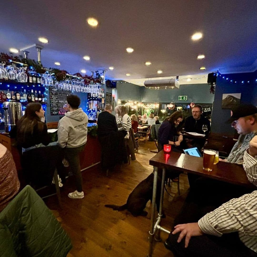 A cozy interior of The Foundation bar, featuring patrons enjoying drinks and conversation. The space is warmly lit with a mix of tables and bar seating. A well-stocked bar is visible in the background, adorned with glassware and festive decorations. A dog rests on the floor, adding to the relaxed atmosphere.