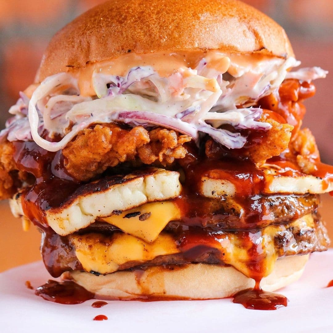 Almost Famous_Mouthwatering Stacked Burger Cheesy_BBQ Sauce Mouthwatering stacked burger from Almost Famous featuring layers of beef patties, cheese, crispy chicken, coleslaw, and sauces.