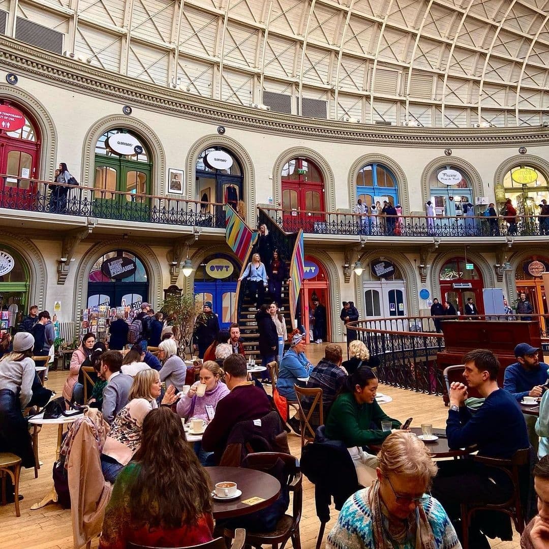 Bruschetta_Victorian_Architecture_Busy_Cafe_Gathering Visitors enjoying the vibrant atmosphere inside the Bruschetta location, with its elegant interior featuring a glass ceiling, arched doorways, and a grand staircase, as people dine and socialize.