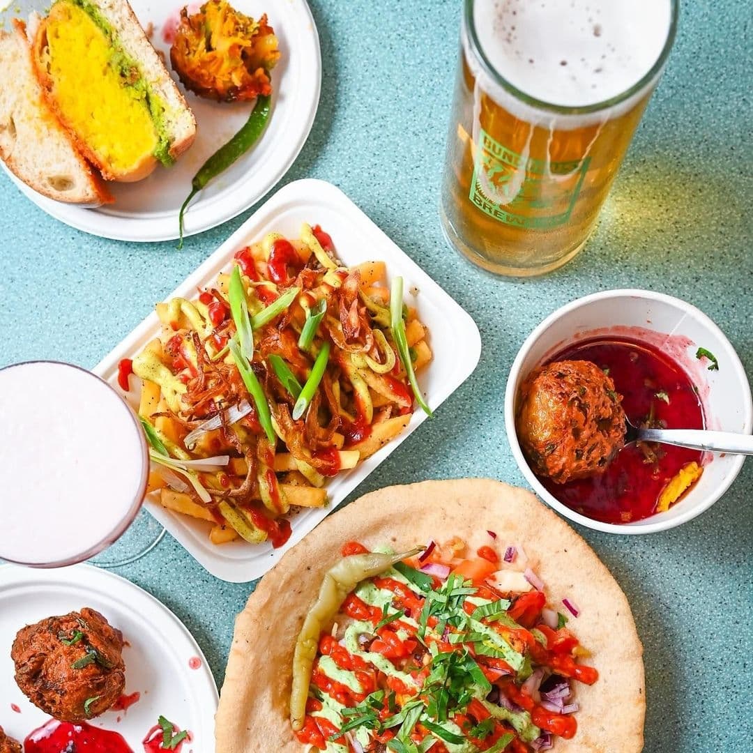 Bundobust_Vibrant_Indian_Cuisine_Colorful_Plates_Craft_Beer Assorted Indian street food dishes and a pint of beer on a textured turquoise table at Bundobust. Visible are a sandwich with vibrant fillings, a bowl of fries with toppings, a round bread with fresh herbs and vegetables, and a deep red sauce with a ball-shaped item, suggesting a flavorful dining experience.