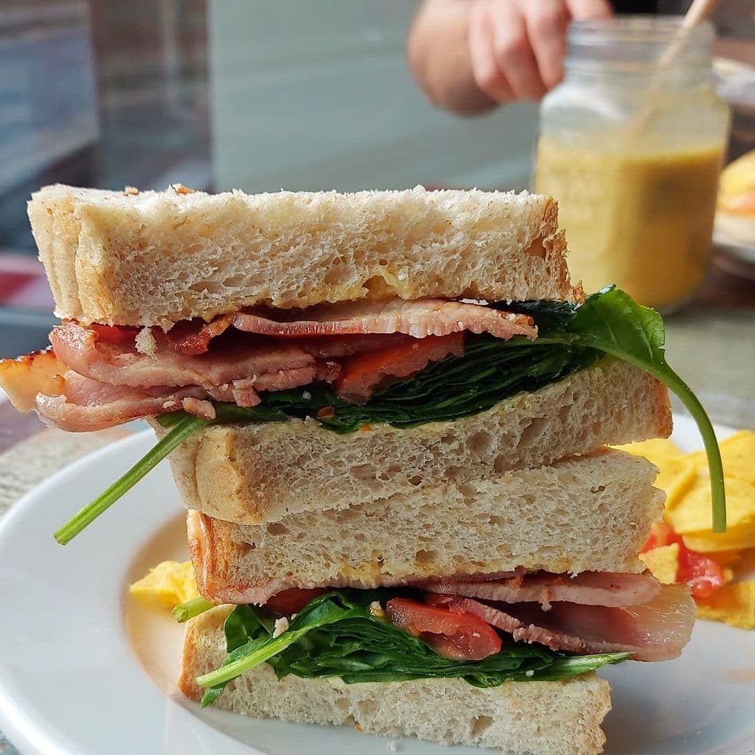 Cha Lounge Delicious BLT Sandwich Crispy Bacon Spinach Close-up of a freshly prepared BLT sandwich with crispy bacon, lettuce, and tomato slices, served on a white plate at Cha Lounge. A jar of sauce and a side of chips are visible in the background.