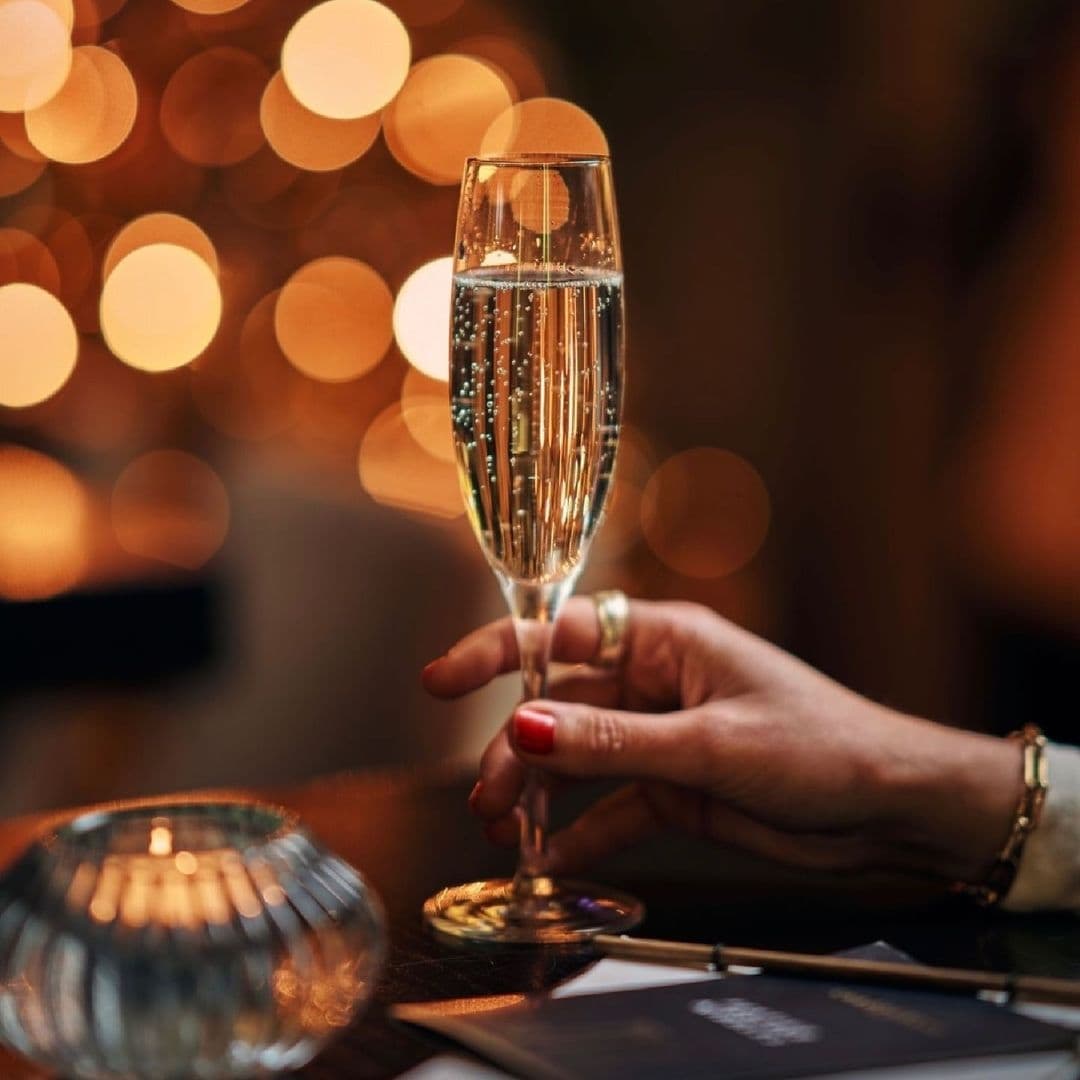 Chaophraya Elegant Champagne Celebration Bokeh Lights Close-up of a person's hand holding a glass of sparkling champagne at Chaophraya, with warm bokeh lights in the background.