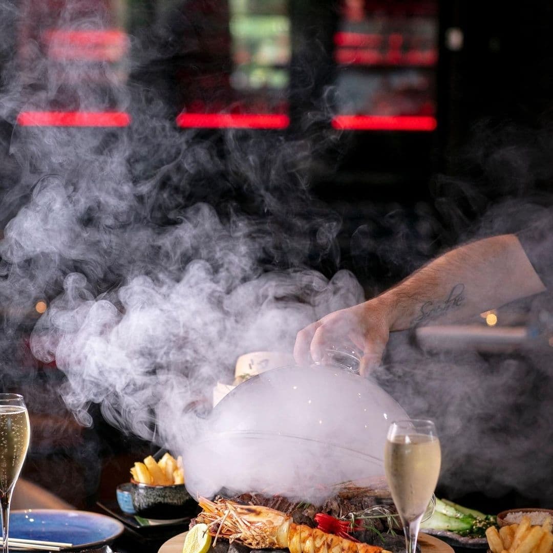 Chino Latino Smoky Culinary Presentation Seafood Dining Alt text: "A tantalizing culinary presentation at Chino Latino with a hand lifting a cloche to reveal a steaming dish, accompanied by a glass of champagne and an array of gourmet sides."