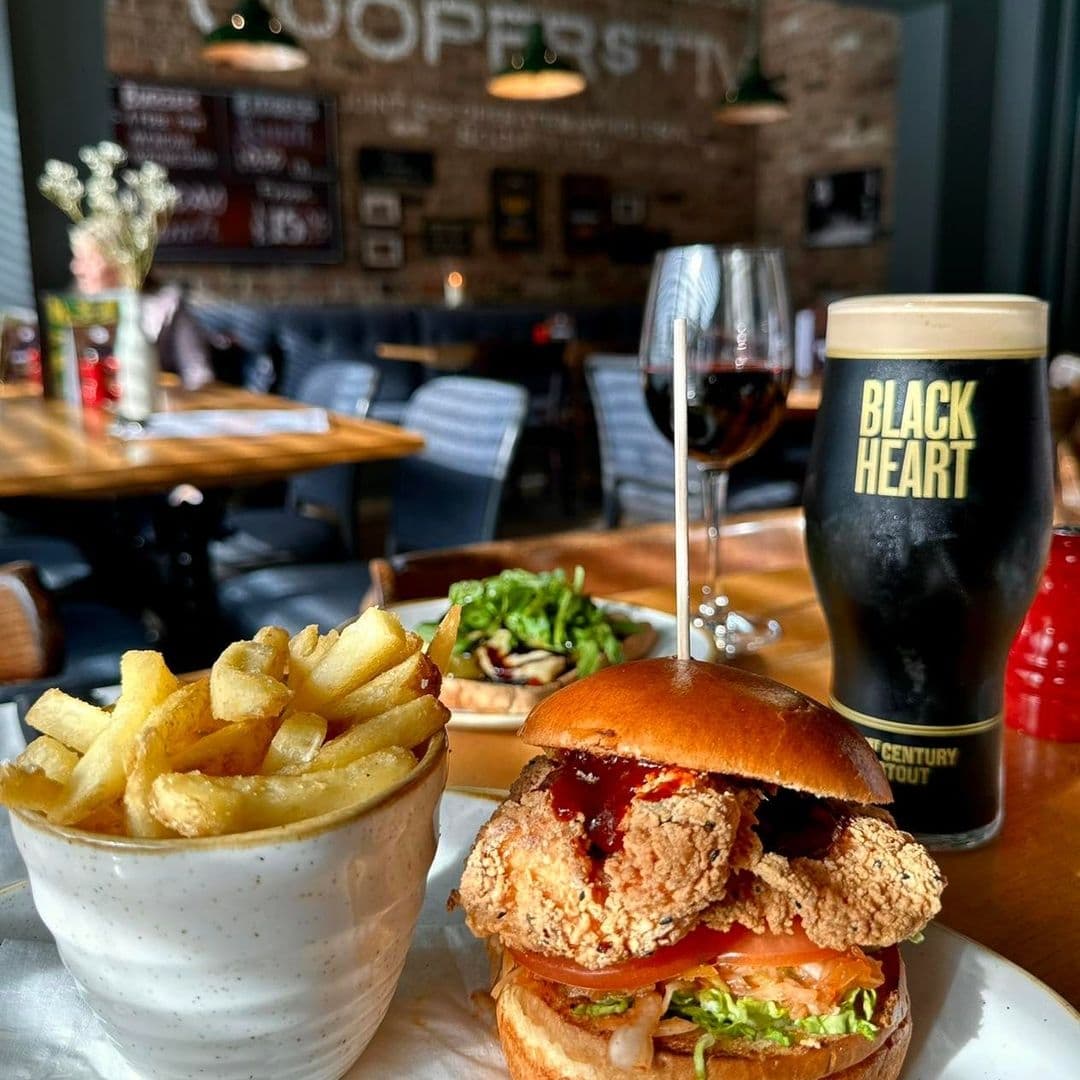 Coopers Bar and Brasserie Gourmet Burger Craft Beer Interior View Cozy dining atmosphere at Coopers Bar and Brasserie featuring a gourmet burger with fries, a pint of stout, and a glass of red wine on a wooden table with the restaurant's interior in the background.