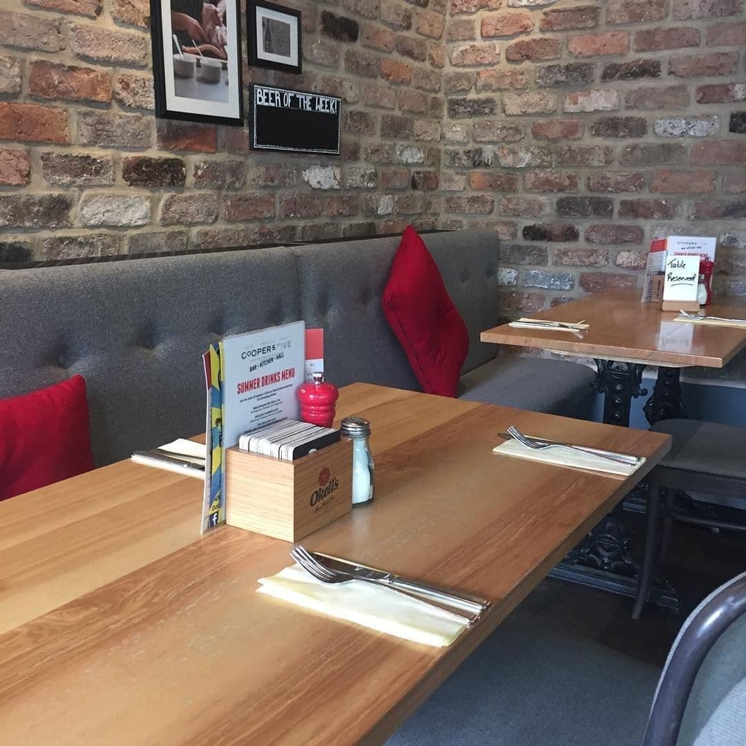 Coopers Bar and Brasserie Inviting Interior Brick Walls Wooden Tables Interior of Coopers Bar and Brasserie showing wooden tables with place settings, a grey upholstered bench, red cushions, and exposed brick walls decorated with framed pictures and a 'Beer of the Week' sign.