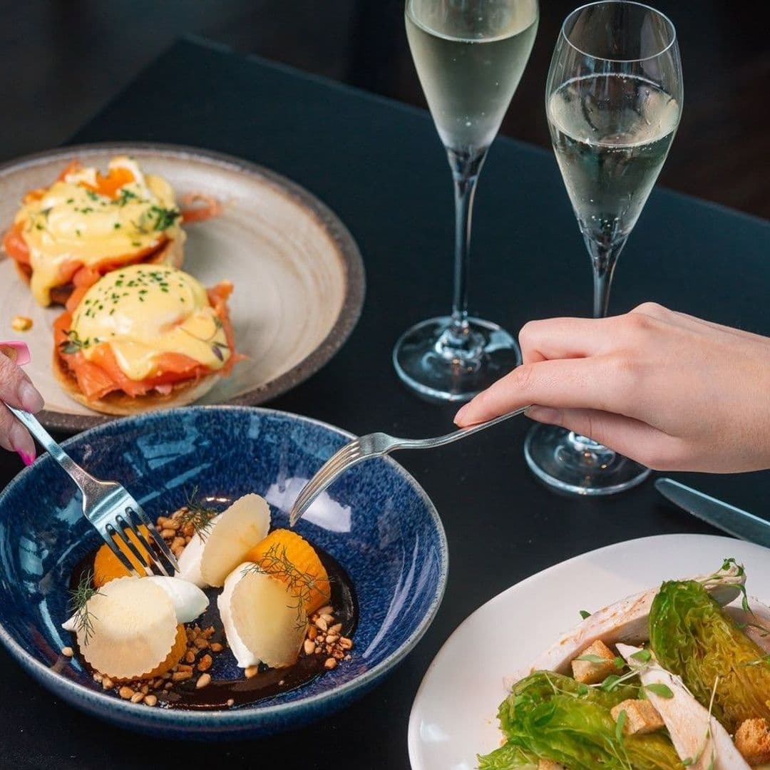 Crafthouse Elegant Dining Experience Champagne Brunch Elegant dining experience at Crafthouse featuring a close-up of gourmet dishes with a person enjoying a meal. Visible are poached eggs on toast, a salad, a dessert with sorbet and chocolate sauce, and two glasses of sparkling wine.
