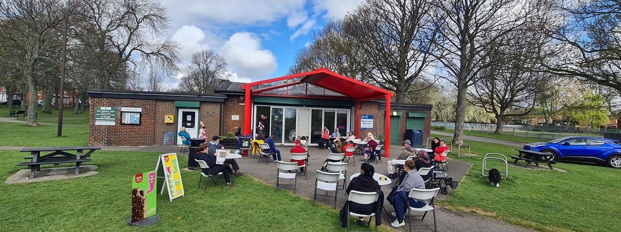 Visitors enjoying outdoor seating at Cross Flatts Park Cafe, with a red canopy, surrounded by greenery and a clear sky.
